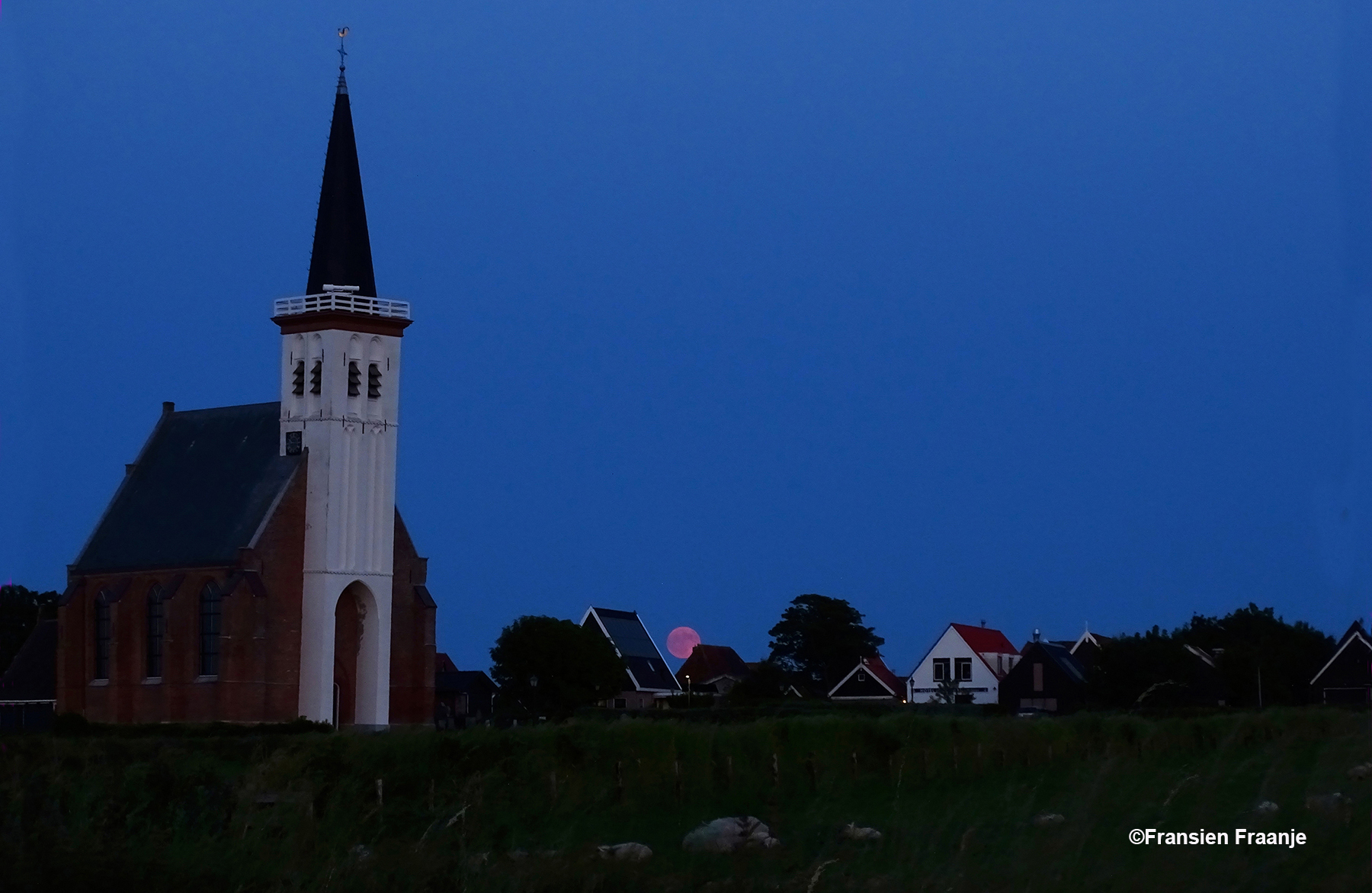 Als wij later terugrijden, zien we tussen de huizen naast het kerkje van Den Hoorn de opkomende maan verschijnen. Onder in beeld zijn nog vaaf de schapen te herkennen in de weide bij het kerkje. - Foto: ©Fransien Fraanje